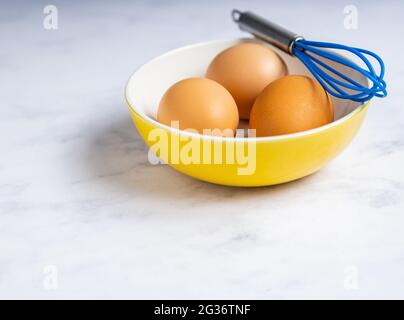 Three brown eggs in a yellow bowl with a blue whisk isolated on a white background. Stock Photo