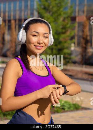 Vertical photo of a young asian woman checks her pulse during the race, smiling and looking at camera. The concept of modern technologies in sports. Stock Photo