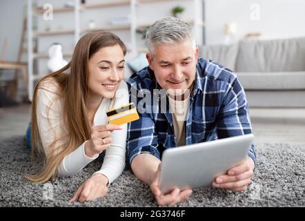 Cheerful mature couple lying on floor with tablet computer, holding credit card, making payment together in online store Stock Photo
