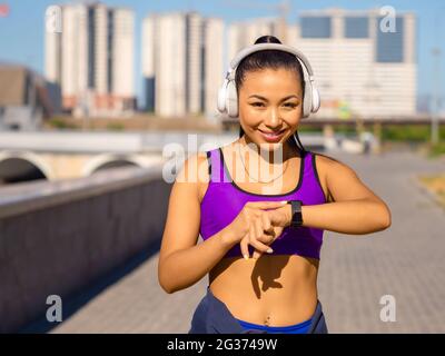 Photo of a young asian woman in white headphones checks her pulse with smartwatch during the race, smiling and looking at camera. Concept of modern te Stock Photo