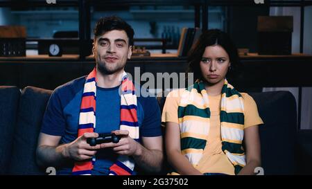 KYIV, UKRAINE - JUNE 09, 2021: cheerful man in striped scarf holding joystick and playing video game near offended girlfriend Stock Photo