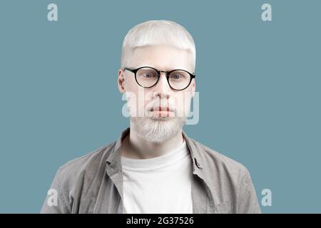 Portrait of serious albino man with pale skin and blue eyes, wearing eyeglasses against turquoise studio background Stock Photo