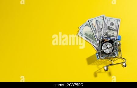 paper bills of one hundred US dollars in a miniature metal cart and a black alarm clock on a yellow background, top view. Time to make money concept, Stock Photo