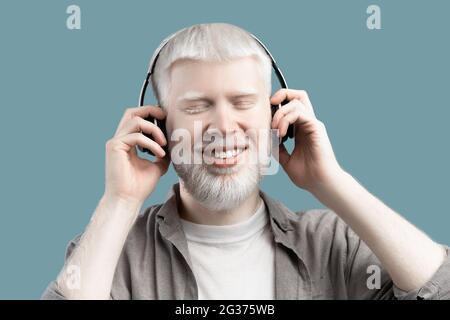 Happy albino man listening music in wireless headphones and touching them on turquoise studio background Stock Photo