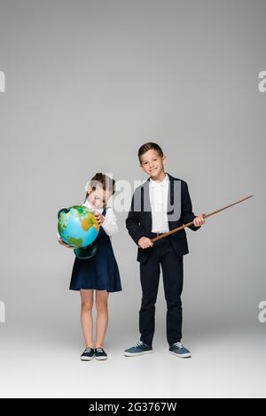 cheerful schoolboy holding pointing stick near sad girl in dress with globe on grey Stock Photo