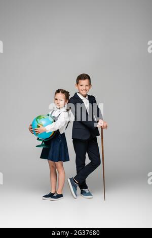 smiling schoolboy holding pointing stick near girl in dress with globe on grey Stock Photo