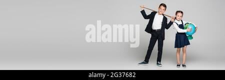happy schoolboy holding pointing stick near girl in dress with globe on grey, banner Stock Photo