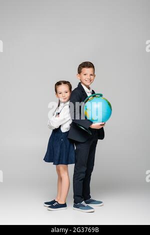 happy schoolboy holding globe while standing with girl in dress on grey Stock Photo