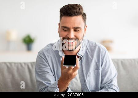 Happy handsome man recording voice message on smartphone or using virtual assistant while resting on sofa at home Stock Photo
