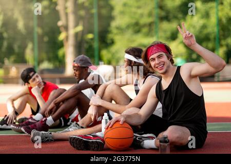 Happy young basketball player and his multinational team having rest at outdoor arena, copy space Stock Photo