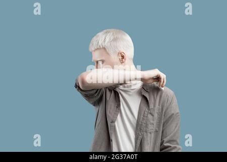 Stop virus and germs spreading concept. Albino man coughing in his elbow, sneezing in correct way, turquoise backgound Stock Photo