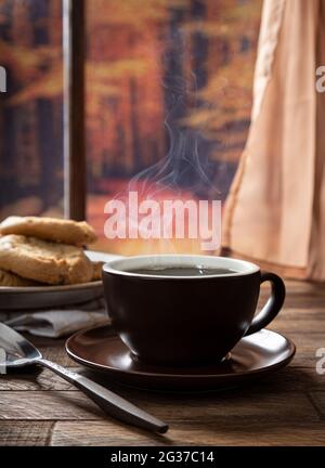 Hot cup of coffee on a wooden table by a window with autumn background Stock Photo