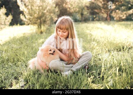 Funny smiling kid girl 6-7 year old playing with spitz dog sitting in green grass in park lawn outdoors. Togetherness. Friendship. Summer season. Stock Photo