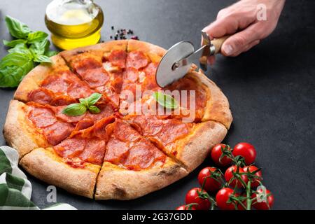 Hand slicing pepperoni pizza on black concrete background. Tasty hot italian pizza cut into slices Stock Photo
