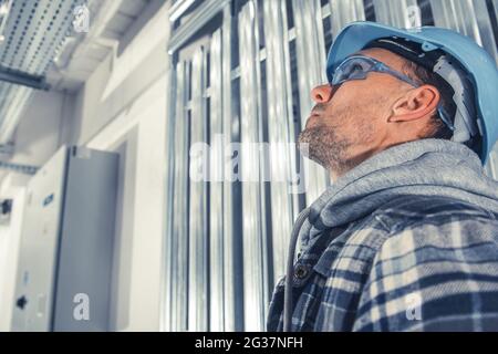 Caucasian Construction Engineer in Blue Hard Hat and Safety Glasses Looking Up. Construction Site Safety. Stock Photo
