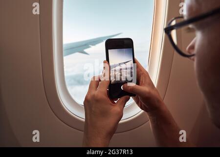 Passenger photographing through airplane window. Man using smart phone during flight above cloouds. Themes travel, connection and memories. Stock Photo