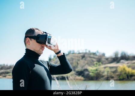 Virtual reality vs real life. Future, Modern technologies. Man in VR virtual reality glasses on the nature background by the river in sun day. Stock Photo