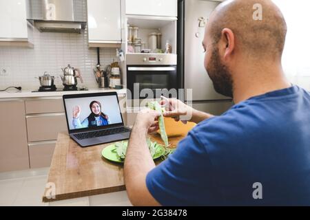 Man chatting with online video while preparing food in kitchen. Man and woman communicating via videoconference while cooking, not neglecting their wo Stock Photo