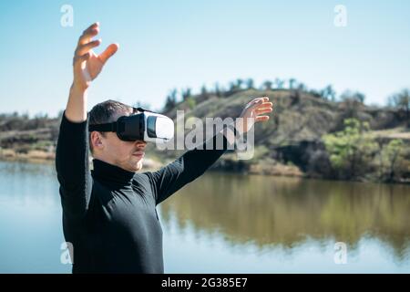 Virtual reality vs real life. Future, Modern technologies. Man in VR virtual reality glasses on the nature background by the river in sun day. Stock Photo