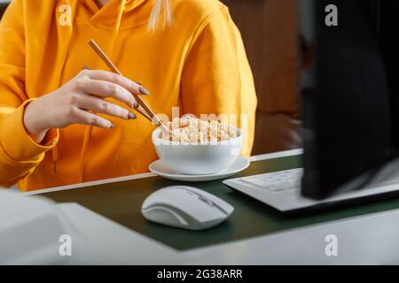 Woman eats noodles with chopsticks behind computer at home workplace, home office during video game or watching movie or education process. Fast junk Stock Photo