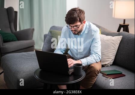 Hard work with the help of a young man's laptop, he sits on the couch, makes a report and drinks an invigorating coffee. Stock Photo