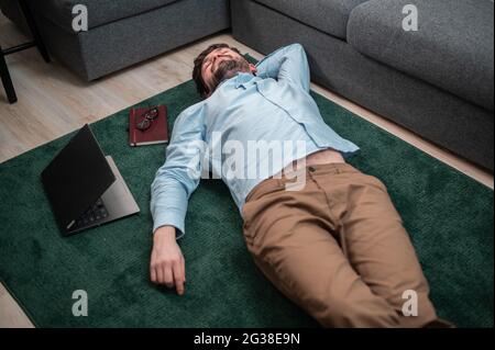 Young cute office worker lies on the floor mat next to the sofa, he is tired and asleep, top view. Stock Photo