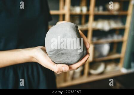 Female hand holding piece of gray clay. Prepare to work in pottery workshop. Shelves with ready goods as background. Stock Photo