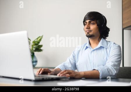 Smiling indian businessman wearing headset is using laptop computer for online communication with customers or colleagues sitting in the modern office Stock Photo