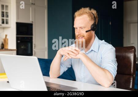 Smiling redhead male freelancer wearing headset and smart casual wear using laptop for connection with colleagues or customers, talking online sitting in the home office Stock Photo