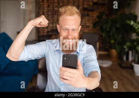 Happy excited redhead bearded hipster guy holding smartphone and looks at the screen, showing triumph gesture and screaming yes, overjoyed lucky young bearded man raising fist up Stock Photo