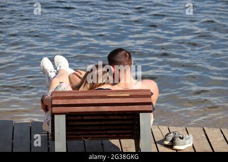 Young couple hugging in a wooden deck chair on a beach. Romantic holiday, summer vacation on a sea or river coast Stock Photo