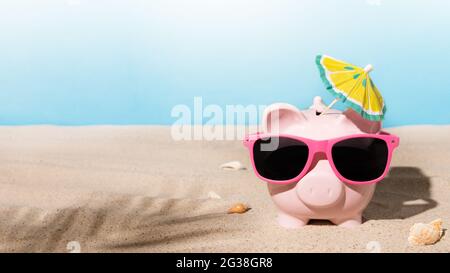 Piggy bank with sunglasses and sunshade umbrella on a sandy beach. Savings for summer vacation or summer sale concept. Stock Photo