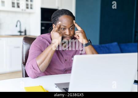 Concentrated African male freelancer using laptop for remote work at home, dark skinned man absorbed with reflections, solving difficult business tasks, staring at the screen focused Stock Photo