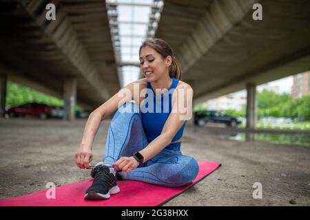 Young athlete sitting on pink mat in parking lot outdoors. Fit woman smiling tying shoelace Stock Photo