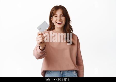 Portrait of young woman laughing and smiling, showing credit card, ready to waste money, paying contactless, recommend bank, standing over white Stock Photo