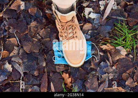 A used protective medical blue mask was stepped on with a brown boot on the fall foliage Stock Photo