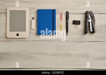 An electronic book, flash memory, notebook, pen, binder and stapler lie on a beige wooden table. There's an empty space at the bottom of the table Stock Photo