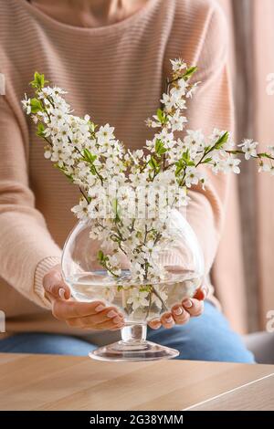 Woman with vase of beautiful blooming branches on table in room Stock Photo