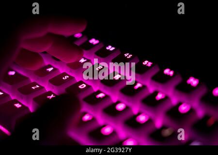 perspective side view detail closeup of male fingers typing on illuminated mechanical gaming RGB keyboard with keycaps lighting up in magenta red colo Stock Photo