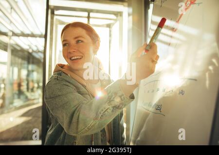 Female student pointing at whiteboard and smiling. Happy young female showing whiteboard in university. Stock Photo