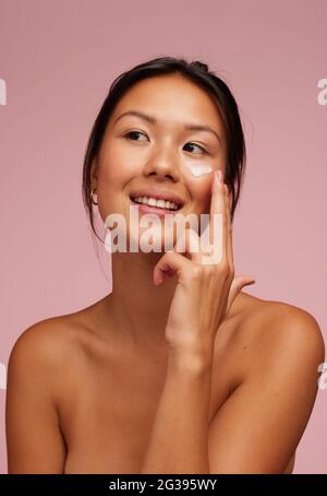 Portrait of a beautiful woman applying moisturizer cream on her face. Woman using beauty product on her facial skin and looking away. Stock Photo