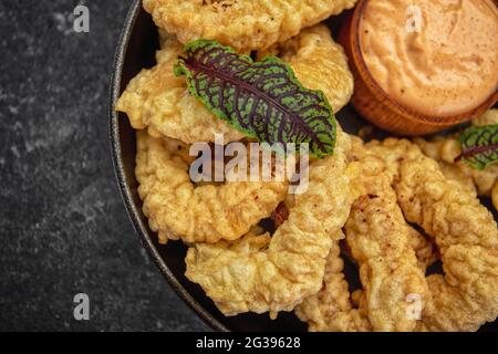 Golden rings of squid fries in a round frying pan with sauce, herbs and lemon on a dark background without decorations Stock Photo