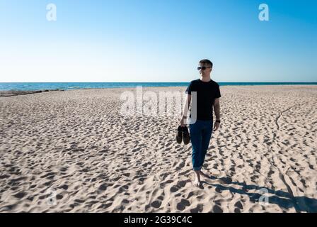 Man in casual wear, glasses stands on beach and holds shoes in hand Stock Photo