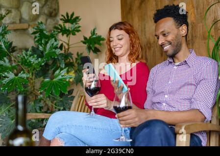 multiethic couple enjoying a glass of wine together outdoor sitting on bench holding a smartphone. diverse happy friends having fun drinking alcohol Stock Photo