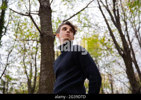 A man stands in an autumn forest, wearing a black wool sweater with a high collar and looking around.Thoughtful young male in a wood. Stock Photo