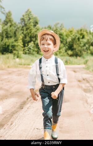 boy in a hat runs across the field. Little farmer on a summer field, happy and cute little boy in a straw hat. portrait of a village boy. boy in rubbe Stock Photo