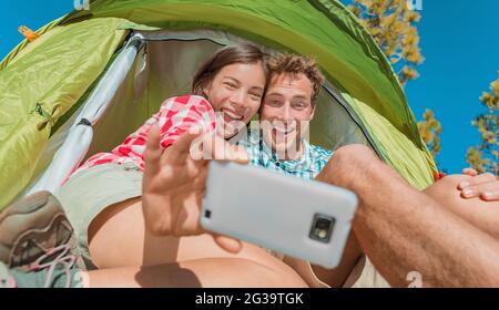 Camping selfie fun couple tourists holding phone taking self portrait photos in tent during summer vacation travel. Asian girl, Caucasian man laughing Stock Photo