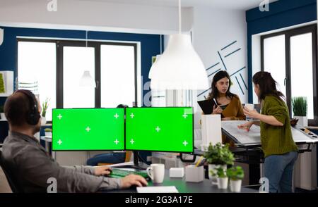 Employee with headphones using dual monitror setup with green screen, chroma key mock up isolated display sitting in video production studio. Man ditor processing film montage on pc in creative agency Stock Photo