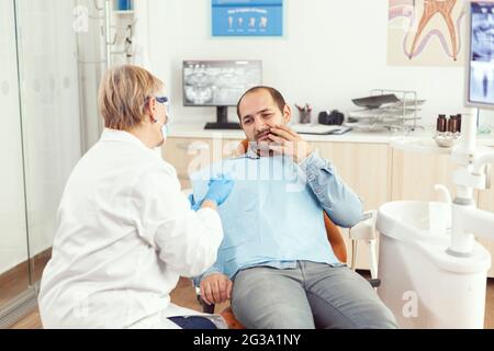 Sick man complaining about tooth pain discussing with stomatologist senior woman about stomatology treatment. Orthodontic doctor preparing for dentistry surgery during medical appointment Stock Photo