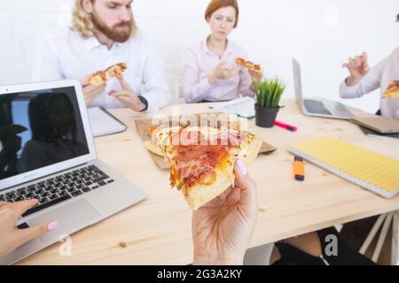 Group of diverse business people eating pizza at break while working together in an office sitting around desk Stock Photo